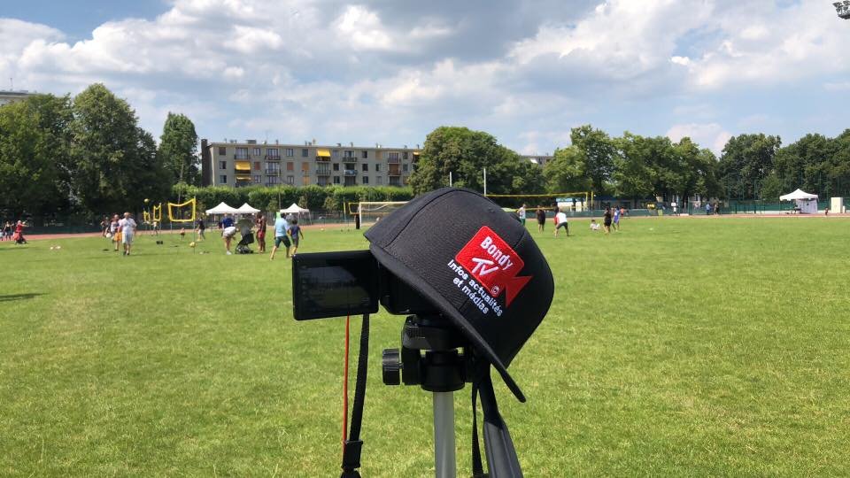 Une caméra sur trépied et une casquette Bondy TV, devant un terrain de sport où des habitants se retrouvent par beau temps